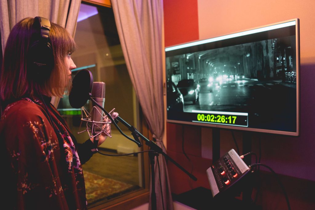 A woman recording a voice over in a vocal booth with a tv Infront of her. ADR